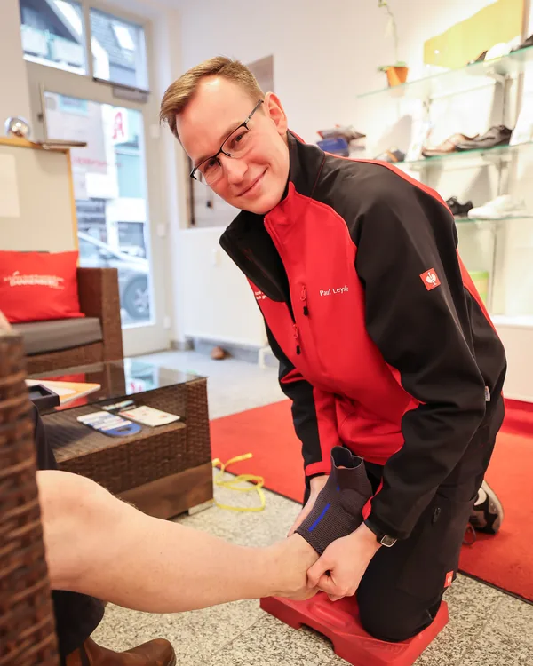 Paul Leyske kniet im Laden, legt einem Kunden eine Bandage am Sprunggelenk an und lächelt in die Kamera
